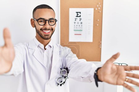 Photo for African american optician man standing by eyesight test looking at the camera smiling with open arms for hug. cheerful expression embracing happiness. - Royalty Free Image