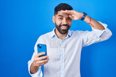 Photo for Hispanic man with beard using smartphone typing message very happy and smiling looking far away with hand over head. searching concept. - Royalty Free Image