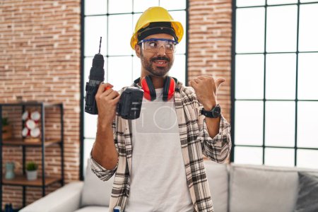 Téléchargez les photos : Jeune homme hispanique avec barbe travaillant à la rénovation de la maison pointant le pouce vers le côté souriant heureux avec la bouche ouverte - en image libre de droit