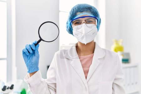 Téléchargez les photos : Young latin woman wearing scientist uniform holding magnifying glass at laboratory - en image libre de droit