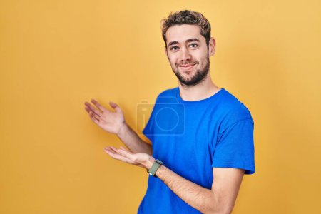 Foto de Hombre hispano con barba de pie sobre fondo amarillo invitando a entrar sonriente natural con la mano abierta - Imagen libre de derechos