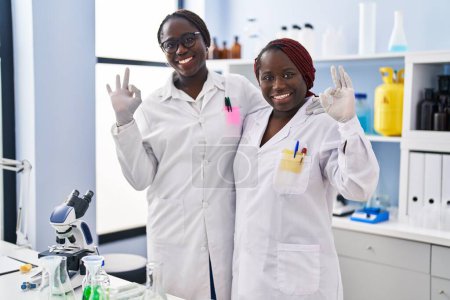 Photo for Two african women working at scientist laboratory doing ok sign with fingers, smiling friendly gesturing excellent symbol - Royalty Free Image