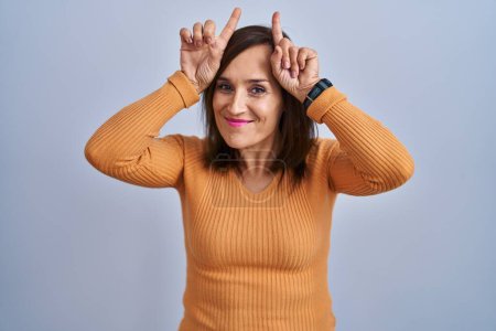 Téléchargez les photos : Femme brune du moyen âge portant un pull orange faisant un geste drôle avec le doigt au-dessus de la tête comme des cornes de taureau - en image libre de droit