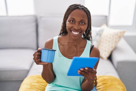 Foto de Mujer afroamericana usando touchpad bebiendo café sentado en el sofá en casa - Imagen libre de derechos