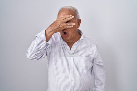 Foto de Senior man with grey hair standing over isolated background peeking in shock covering face and eyes with hand, looking through fingers with embarrassed expression. - Imagen libre de derechos