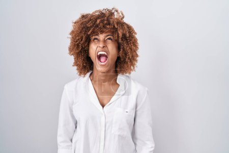 Foto de Young hispanic woman with curly hair standing over white background angry and mad screaming frustrated and furious, shouting with anger. rage and aggressive concept. - Imagen libre de derechos