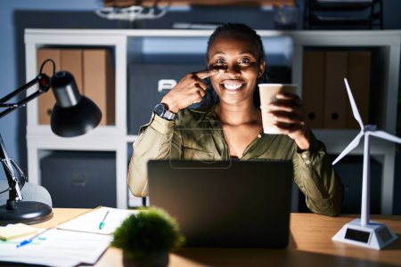 Photo for African woman working using computer laptop at night pointing with hand finger to face and nose, smiling cheerful. beauty concept - Royalty Free Image
