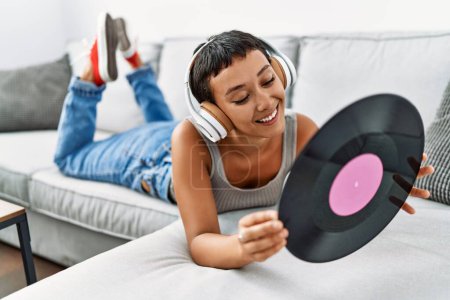 Photo for Young hispanic woman listening to music holding vinyl disc lying on sofa at home - Royalty Free Image