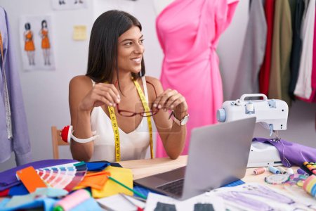 Téléchargez les photos : Jeune belle femme hispanique tailleur souriant confiant assis sur la table au magasin de tailleur - en image libre de droit