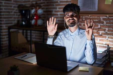 Foto de Joven hombre hispano con barba trabajando en la oficina por la noche mostrando y señalando con los dedos el número nueve mientras sonríe confiado y feliz. - Imagen libre de derechos