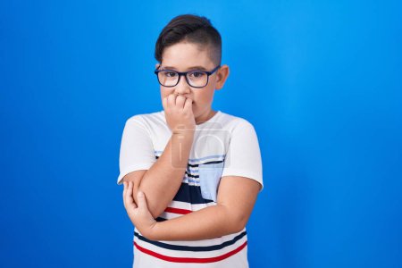 Photo for Young hispanic kid standing over blue background looking stressed and nervous with hands on mouth biting nails. anxiety problem. - Royalty Free Image