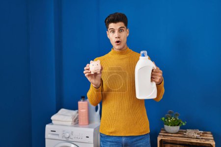 Foto de Joven hombre hispano sosteniendo botella de detergente y alcancía en la cara de choque, mirando escéptico y sarcástico, sorprendido con la boca abierta - Imagen libre de derechos