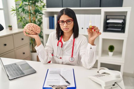Photo for Young hispanic doctor woman holding brain and cbd oil relaxed with serious expression on face. simple and natural looking at the camera. - Royalty Free Image