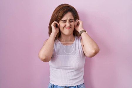 Foto de Mujer morena de pie sobre fondo rosa cubriendo las orejas con los dedos con expresión molesta por el ruido de la música fuerte. concepto de sordo. - Imagen libre de derechos