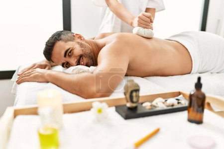 Photo for Young hispanic man having back massage using thai bags at beauty center - Royalty Free Image