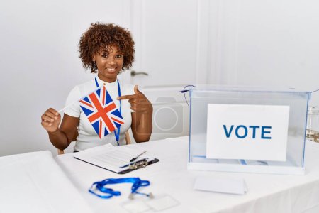 Photo for Young african american woman at political campaign election holding uk flag smiling happy pointing with hand and finger - Royalty Free Image