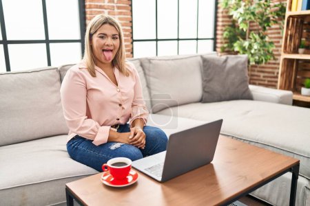 Foto de Young hispanic woman using laptop sitting on the sofa at home sticking tongue out happy with funny expression. emotion concept. - Imagen libre de derechos