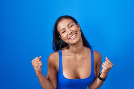 Photo for Hispanic woman standing over blue background very happy and excited doing winner gesture with arms raised, smiling and screaming for success. celebration concept. - Royalty Free Image