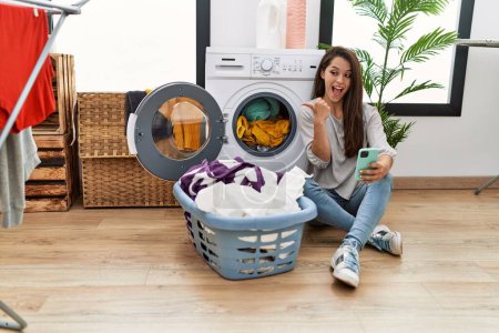 Photo for Young brunette woman doing laundry using smartphone pointing thumb up to the side smiling happy with open mouth - Royalty Free Image