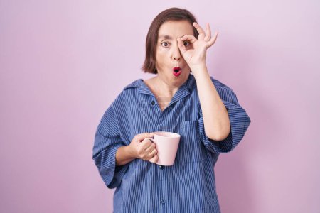 Foto de Mujer hispana de mediana edad bebiendo una taza de café haciendo un gesto bien sorprendido con la cara sorprendida, los ojos mirando a través de los dedos. expresión incrédula. - Imagen libre de derechos