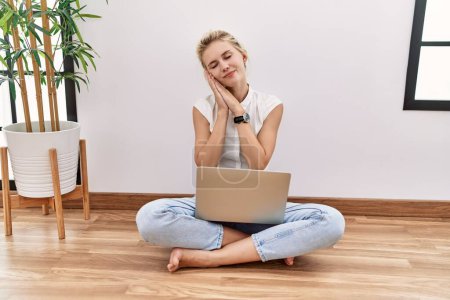 Téléchargez les photos : Jeune femme blonde utilisant un ordinateur portable assis sur le sol du salon endormi fatigué rêvant et posant les mains ensemble tout en souriant les yeux fermés. - en image libre de droit
