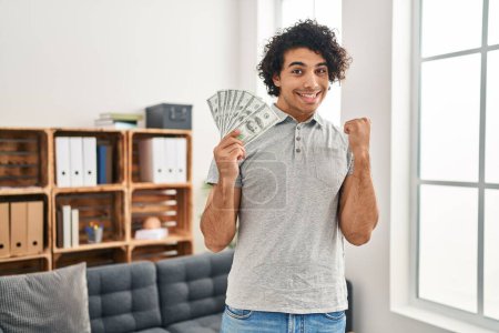 Photo for Hispanic man with curly hair holding 100 dollars banknotes screaming proud, celebrating victory and success very excited with raised arm - Royalty Free Image