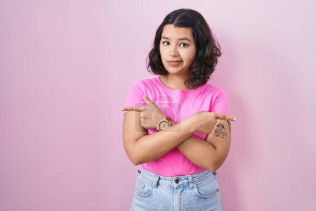 Téléchargez les photos : Jeune femme hispanique debout sur fond rose pointant des deux côtés avec les doigts, direction différente en désaccord - en image libre de droit