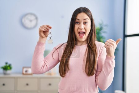 Téléchargez les photos : Jeune femme brune tenant les clés de la nouvelle maison pointant le pouce vers le côté souriant heureux avec bouche ouverte - en image libre de droit