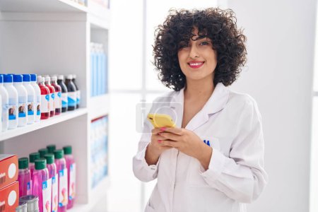 Foto de Joven farmacéutica de Oriente Medio sonriendo confiada usando teléfono inteligente en la farmacia - Imagen libre de derechos
