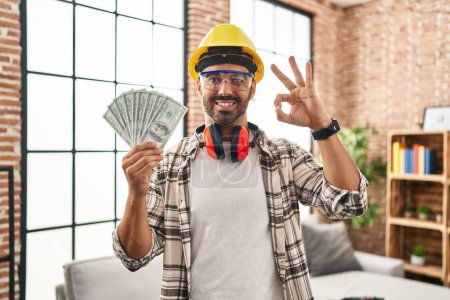 Foto de Joven hombre hispano con barba trabajando en la renovación del hogar sosteniendo dólares haciendo señal de ok con los dedos, sonriendo gesto amistoso excelente símbolo - Imagen libre de derechos
