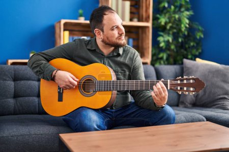Téléchargez les photos : Jeune homme hispanique jouant de la guitare classique assis sur le lit à la maison - en image libre de droit