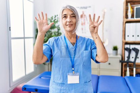 Photo for Middle age grey-haired woman wearing physiotherapist uniform at medical clinic showing and pointing up with fingers number nine while smiling confident and happy. - Royalty Free Image