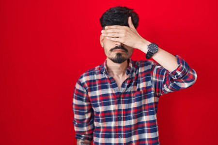 Photo for Young hispanic man with beard standing over red background covering eyes with hand, looking serious and sad. sightless, hiding and rejection concept - Royalty Free Image
