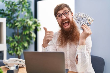 Photo for Caucasian man with long beard working using computer laptop holding dollars pointing thumb up to the side smiling happy with open mouth - Royalty Free Image