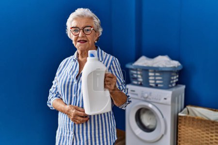 Foto de Mujer mayor con cabello gris sosteniendo botella de detergente que se ve positiva y feliz de pie y sonriendo con una sonrisa confiada mostrando los dientes - Imagen libre de derechos