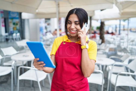 Foto de Joven hermosa mujer árabe camarera hablando en el teléfono inteligente usando touchpad en la terraza de la cafetería - Imagen libre de derechos