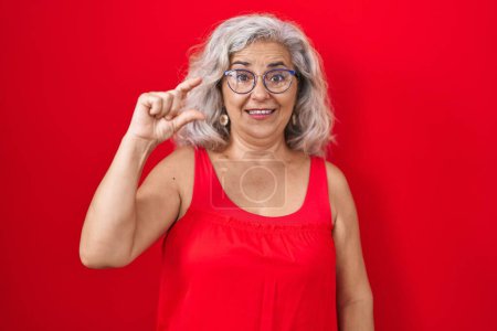 Téléchargez les photos : Femme d'âge moyen avec les cheveux gris debout sur fond rouge geste souriant et confiant avec la main faisant signe de petite taille avec les doigts regardant et la caméra. concept de mesure. - en image libre de droit