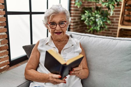Téléchargez les photos : Femme âgée aux cheveux gris lisant une bible assise sur un canapé à la maison - en image libre de droit