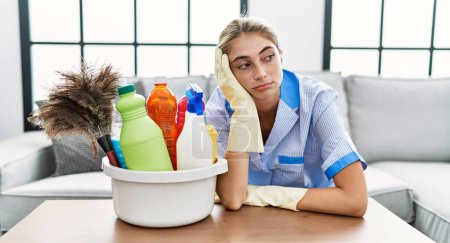 Photo for Young caucasian woman tired wearing cleaner uniform at home - Royalty Free Image