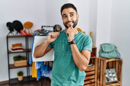 Photo for Young handsome man with beard holding shopping bags at retail shop with hand on chin thinking about question, pensive expression. smiling and thoughtful face. doubt concept. - Royalty Free Image