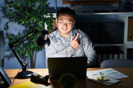 Téléchargez les photos : Jeune homme chinois travaillant à l'aide d'un ordinateur portable la nuit joyeuse avec un sourire du visage pointant avec la main et le doigt sur le côté avec une expression heureuse et naturelle sur le visage - en image libre de droit