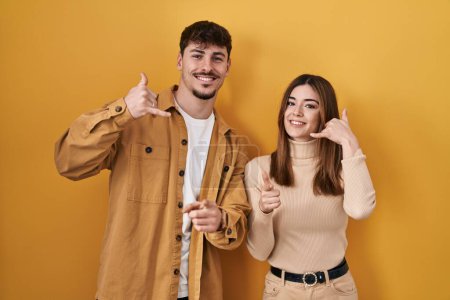 Téléchargez les photos : Jeune couple hispanique debout sur fond jaune souriant faisant parler au téléphone geste et vous pointant du doigt. Appelle-moi. - en image libre de droit