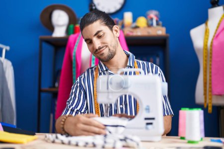 Foto de Joven hombre hispano sastre sonriendo confiado usando máquina de coser en estudio de costura - Imagen libre de derechos