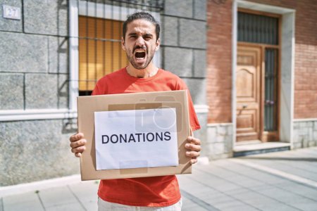 Téléchargez les photos : Young hispanic man holding donations box for charity outdoors angry and mad screaming frustrated and furious, shouting with anger. rage and aggressive concept. - en image libre de droit