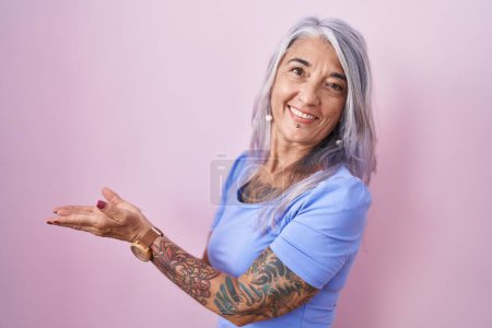 Foto de Mujer de mediana edad con tatuajes de pie sobre fondo rosa señalando a un lado con las manos abiertas palmeras mostrando espacio de copia, presentando anuncio sonriendo emocionado feliz - Imagen libre de derechos