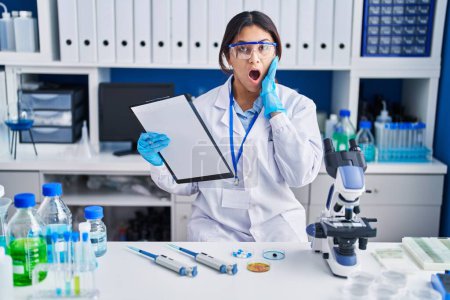 Photo for Hispanic young woman working at scientist laboratory afraid and shocked, surprise and amazed expression with hands on face - Royalty Free Image