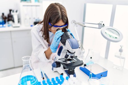 Téléchargez les photos : Jeune fille hispanique regardant à travers le microscope au laboratoire - en image libre de droit