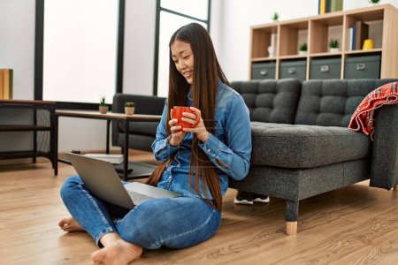 Téléchargez les photos : Jeune fille chinoise en utilisant un ordinateur portable et boire du café assis sur le sol à la maison. - en image libre de droit