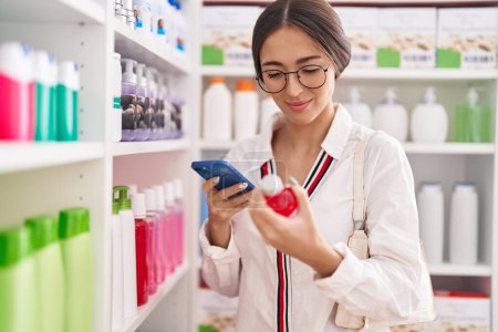 Téléchargez les photos : Jeune belle hispanique femme client en utilisant smartphone tenant bouteille de médicament à la pharmacie - en image libre de droit
