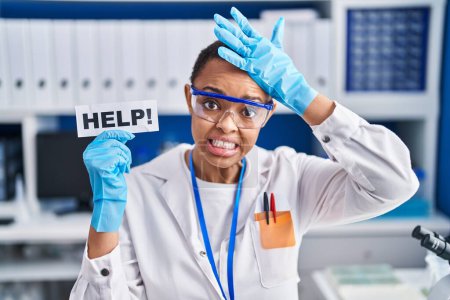 Téléchargez les photos : Belle femme afro-américaine travaillant au laboratoire scientifique demandant de l'aide stressée et frustrée avec la main sur la tête, visage surpris et en colère - en image libre de droit